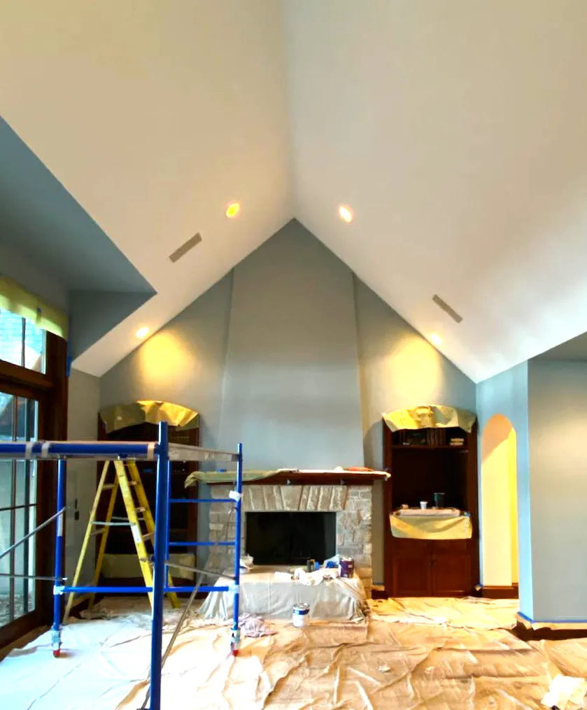 A vaulted living room with a stone fireplace, walls newly painted blue-gray, scaffolding and drop cloths still in place.