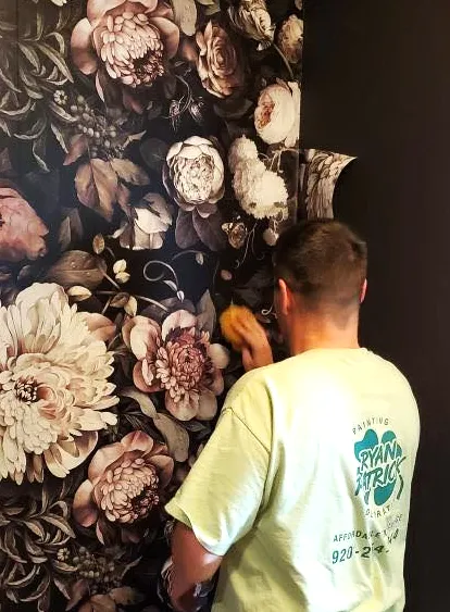 A painter in a RyanPatrick shirt smoothing a dark floral mural wallpaper onto a wall.