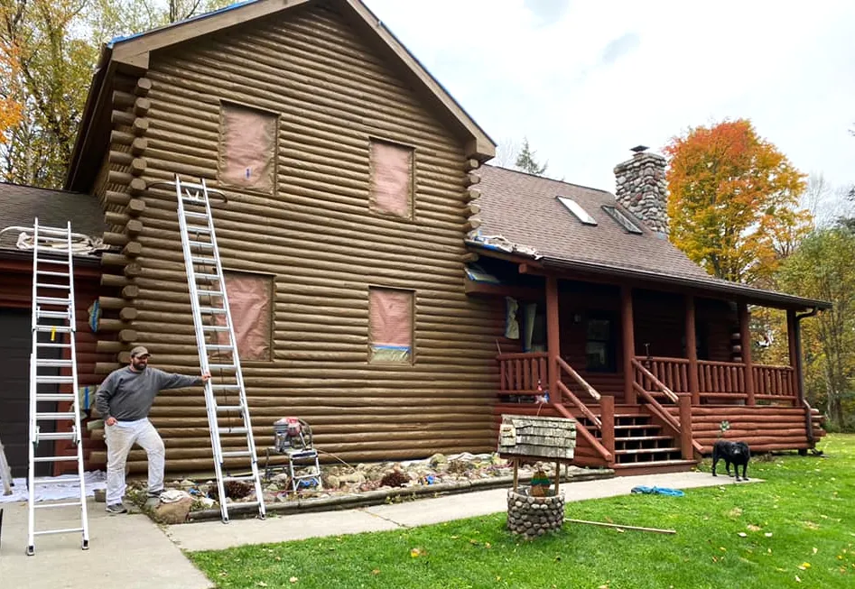 Ryan standing beside ladders at a two-story log home with its windows masked for painting.