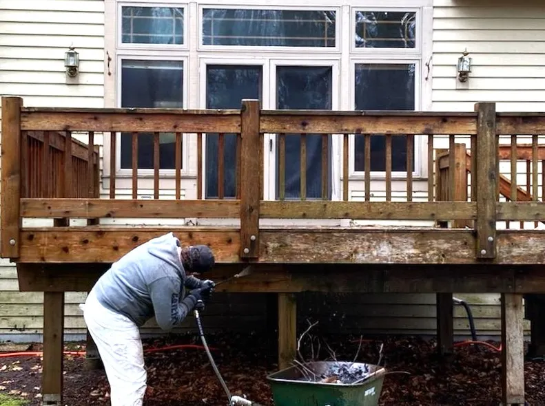A worker pressure washing the weathered wood railing and frame of a raised deck.