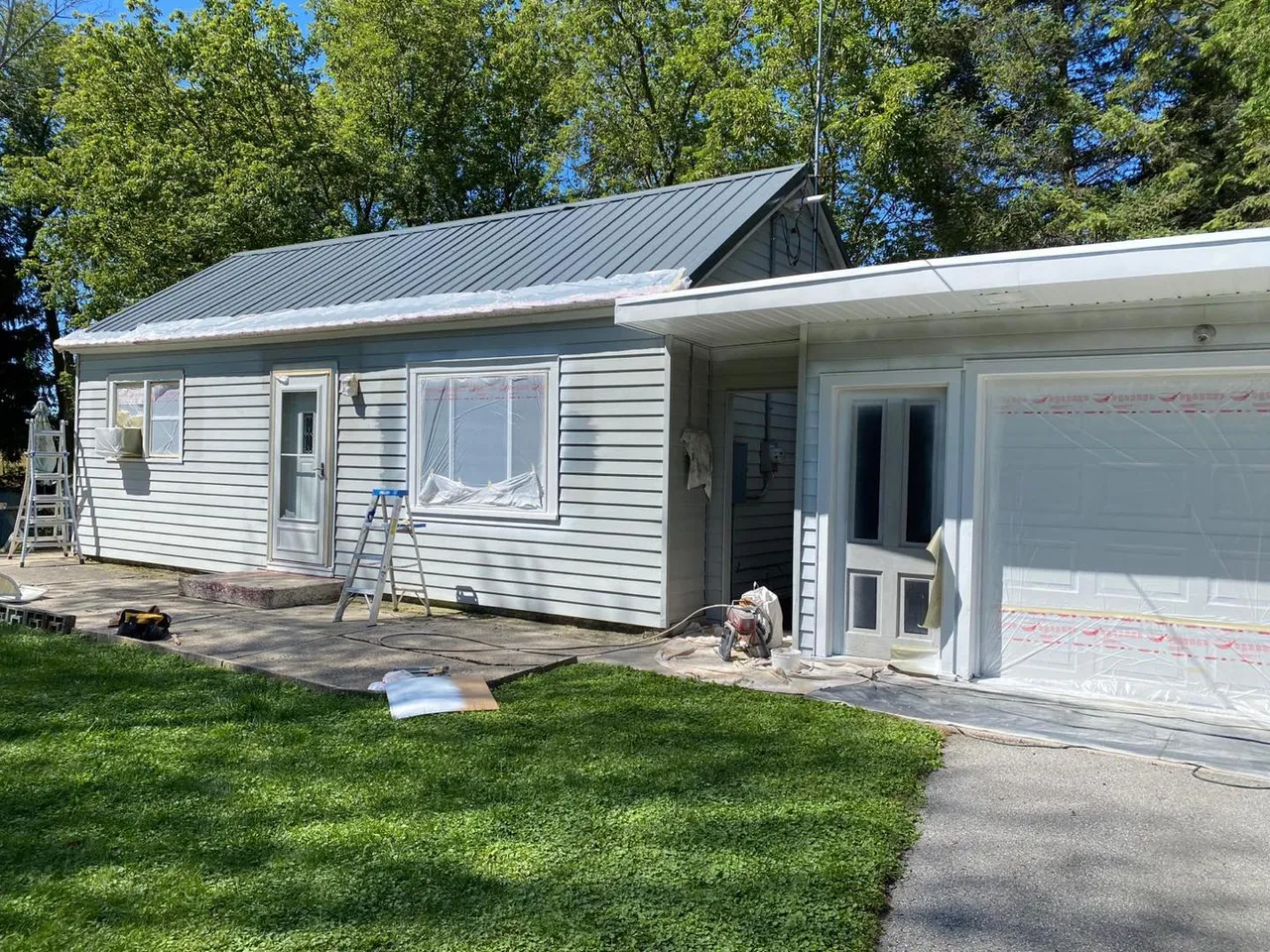 The same ranch house before painting, with pale siding, windows covered in plastic and a paint sprayer on the walk.
