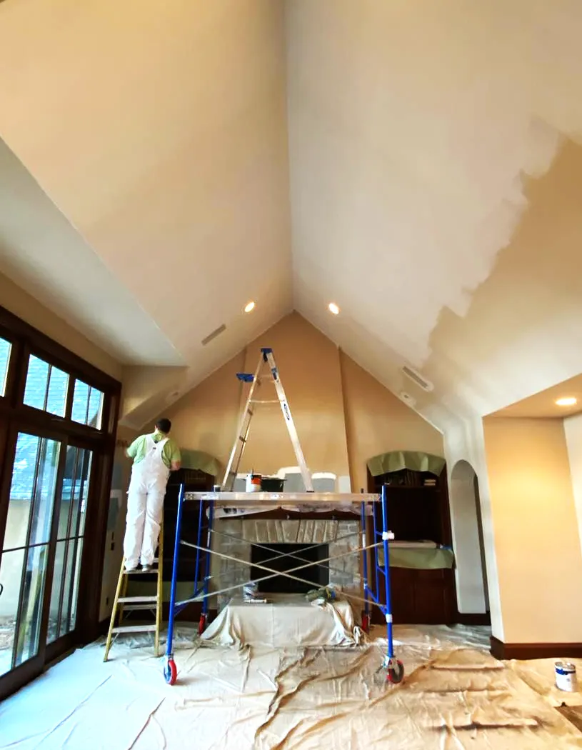 The professionals at RyanPatrick Painting & Decorating in a large room with a tall vaulted ceiling, a fireplace, and large picturesque windows applying primer and a professional finish to the interior of a house.